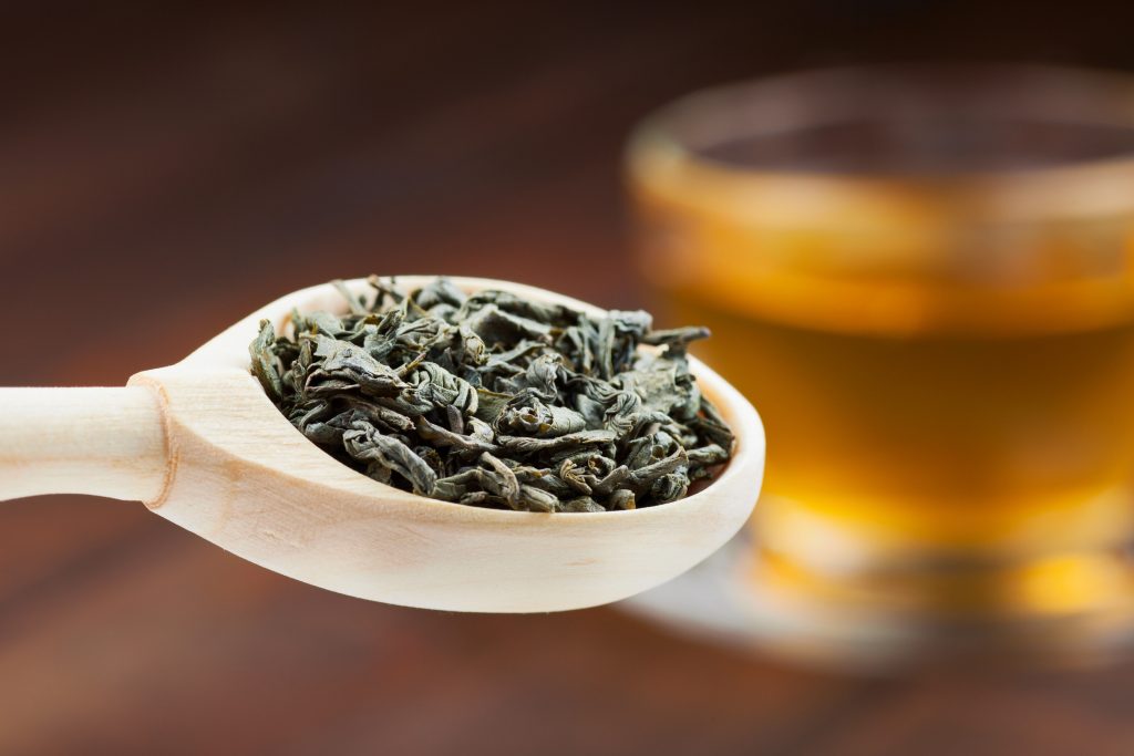green tea leaves in wooden spoon, cup of tea on background